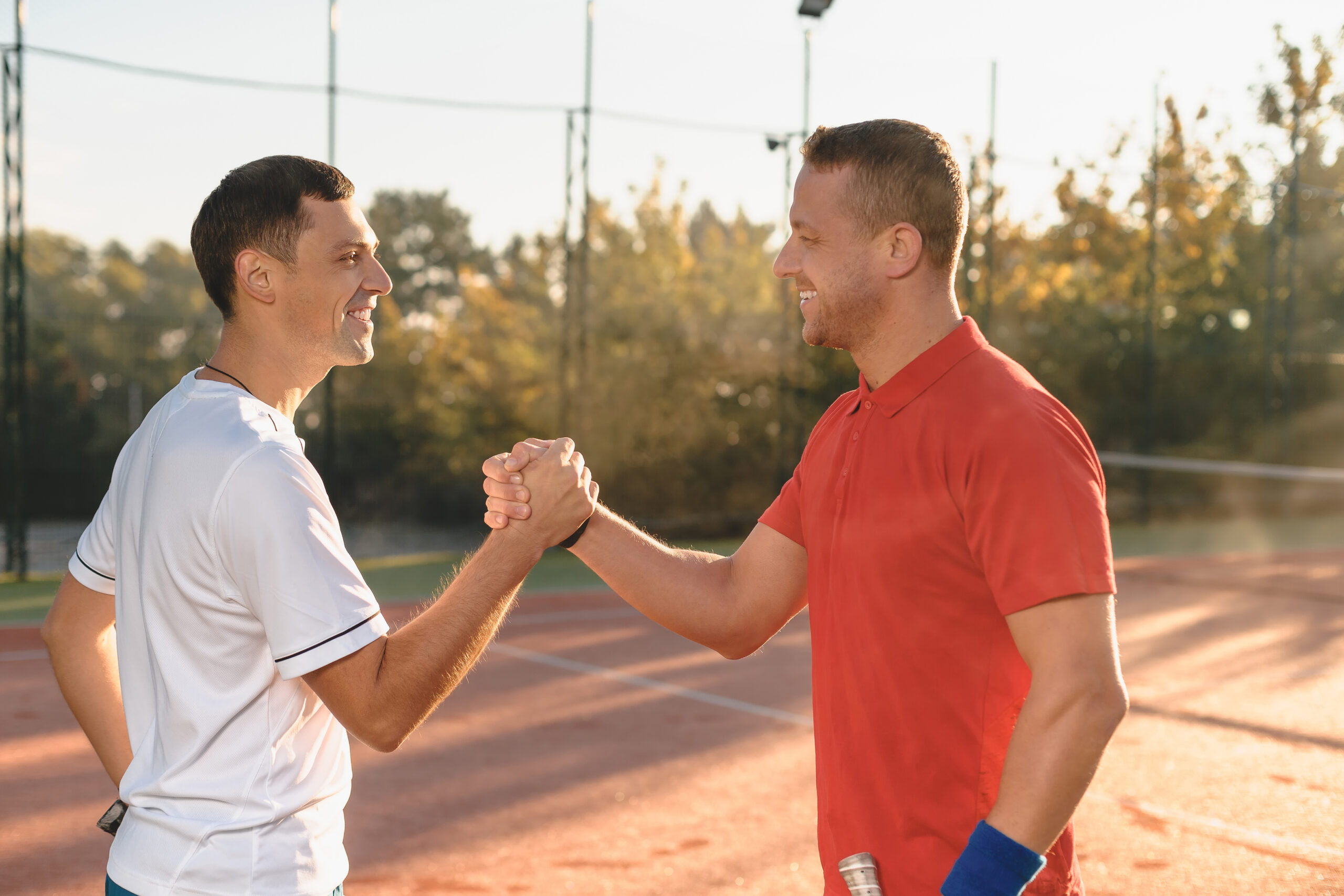 Two determined men are shaking hands smiling, standing in morning sunlight outdoors. Concept sport, health and friendship.