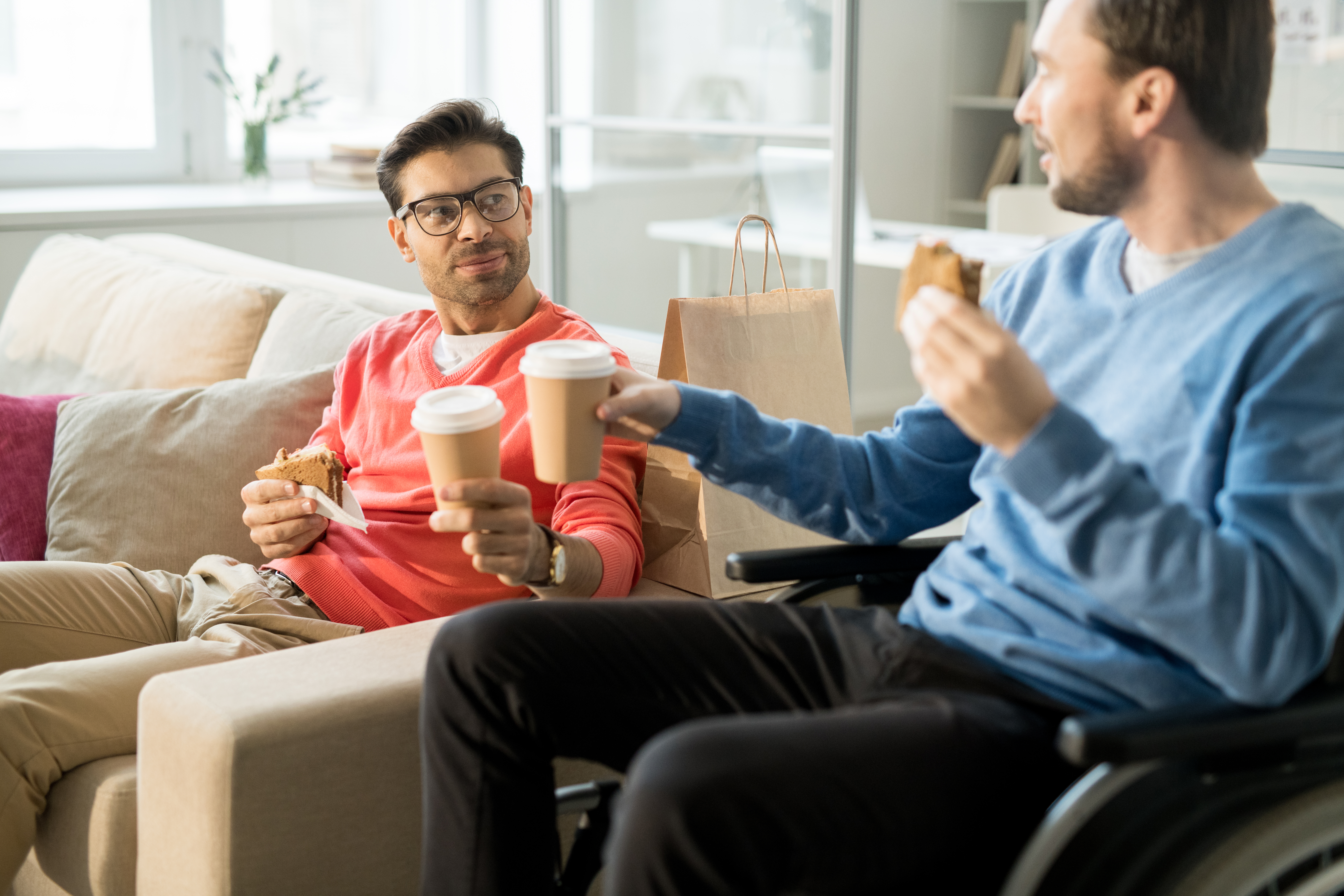 Young businessman in casual clothing sitting on sofa and drinking coffee with his disabled colleague in wheelchair
