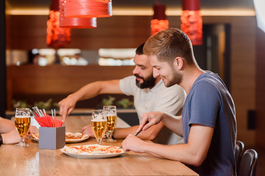Side view of two male friends eating delicious pizza with help of knife and fork. They also have glasses of tasty beer on wooden table. Interior of modern pizzeria or cafe.